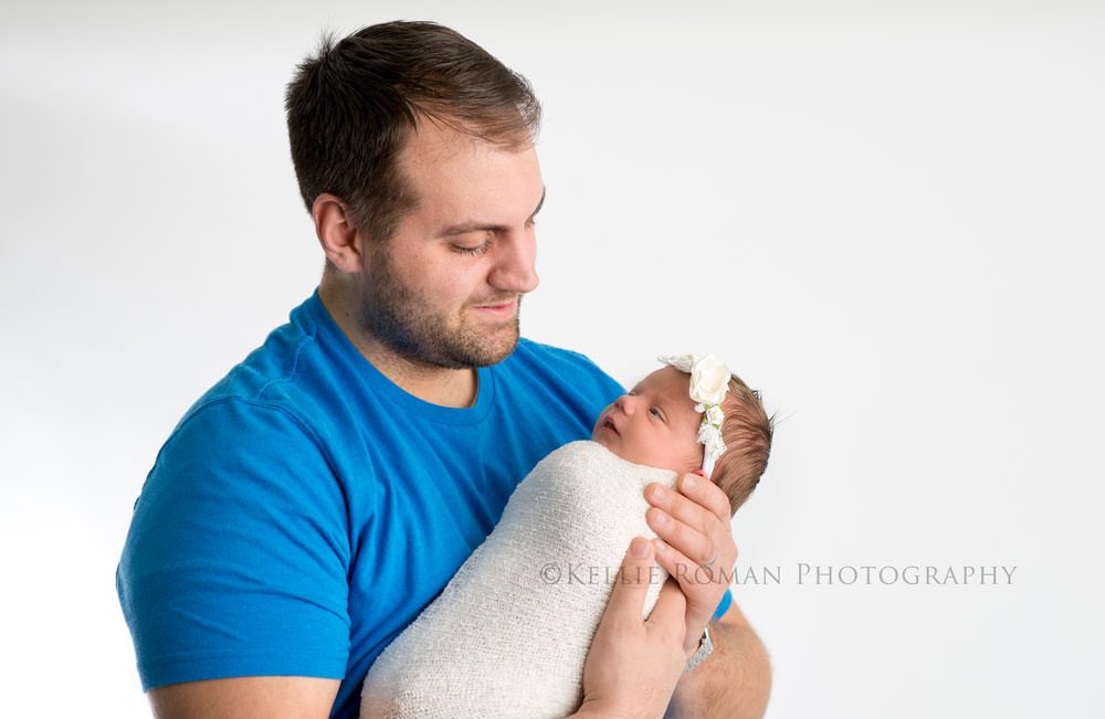 newborn pics milwaukee studio a father who is wearing bright blue shirt and is holding his newborn daughter while looking at her she is sleeping and wrapped in white cloth