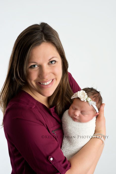 newborn pics milwaukee studio a mom who is holding her baby girl and is looking at the camera mom is wearing a maroon shirt and baby is in a white swaddle with a white headband and is sleeping