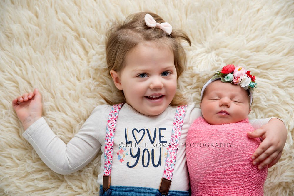 newborn pics milwaukee studio a toddler girl wearing a shirt and suspenders is laying on her back with her newborn sister next to her toddler girl has her arm around the newborn who is in a pink swaddle with a headband