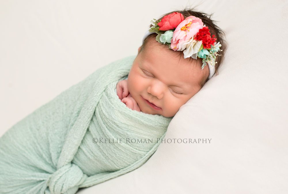 newborn pics milwaukee studio a little newborn girl wrapped in a mint green swaddle with a knot tied in the center she is on top of a cream blanket and is wearing a pink red and teal headband she has both her hands up near her face and is smiling