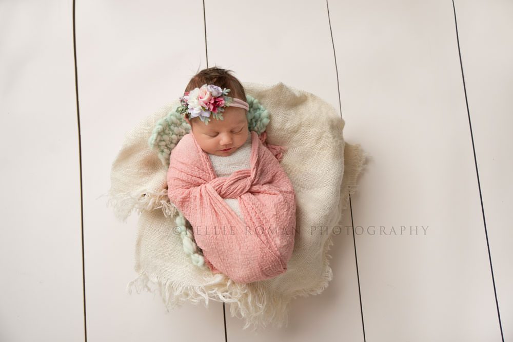 newborn pics milwaukee studio a newborn girl from a downward shot she is in a pink swaddle wrap with a tie in the center and she's laying onto of a teal blanket and white burlap she has a pink and teal floral headband on