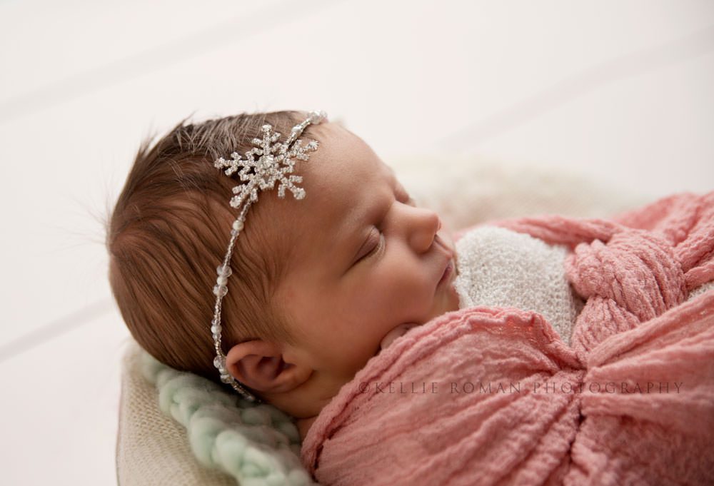 newborn pics milwaukee studio a baby girl wrapped in a pink swaddle wearing a crystal snowflake headband she's laying on her back in a white vintage tub