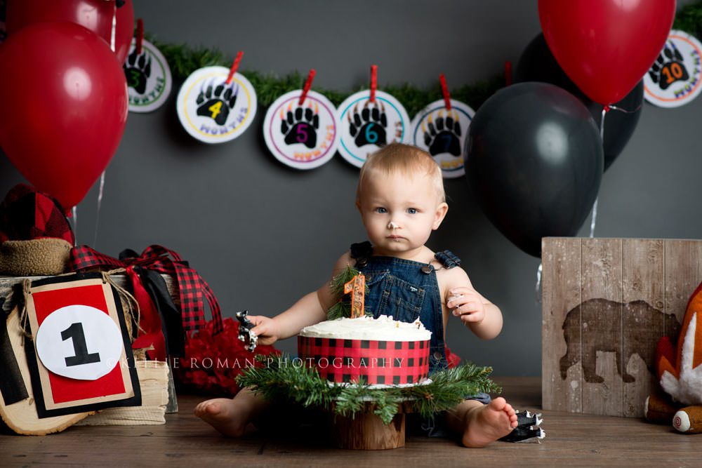 lumberjack cake smash a one year old boy sitting behind a festive cake with the number one on it he has frosting on his nosed and is surrounded by outdoor themed decor
