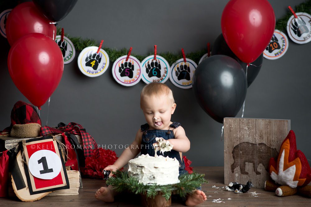 lumberjack cake smash a one year old boy sitting behind a cake with his hand covered in frosting he's licking frosting off his lips he's surrounded by lumberjack themed decor and balloons