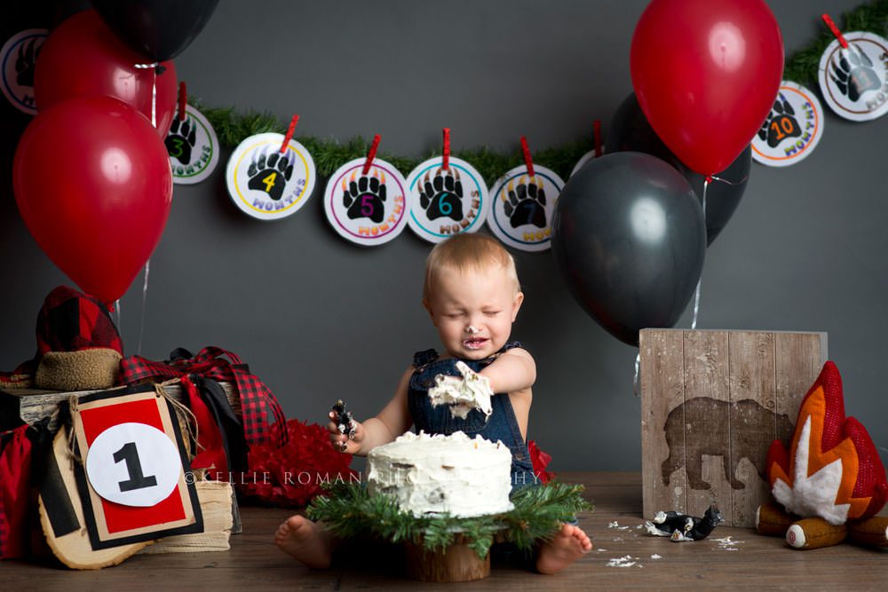 lumberjack cake smash a one year old boy with white frosting all over his hand and he is crying he's surrounded by black and red birthday decor and he's wearing denim overalls