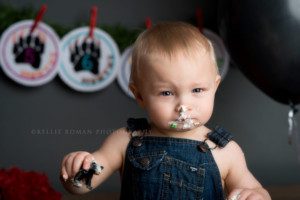 lumberjack cake smash a one year old boy wearing overalls with a smirk on his face he has frosting all over his mouth and nose and is holding onto a frosting covered black bear cake topped