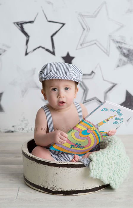 simple cake smash a one year old boy sitting in a vintage white bucket holding a book he has a seersucker romper and hat on he's sitting in front of a black and white star backdrop
