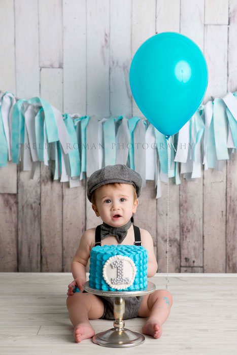 simple cake smash a little boy sitting behind a teal cake with the number one written on it he is wearing a vintage outfit with a wood backdrop and teal and white rag banner
