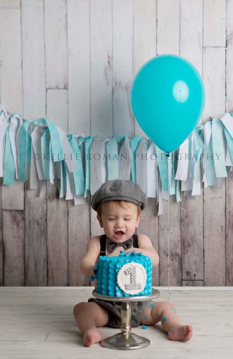 simple cake smash a one year old boy wearing a vintage outfit sitting behind a teal cake on a silver stand there is a distressed wood backdrop with a teal and grey rag banner