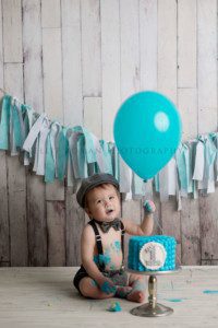 simple cake smash a little boy holding onto a teal colored balloon with a teal cake in front front of him he has a tow tie hat and suspenders on