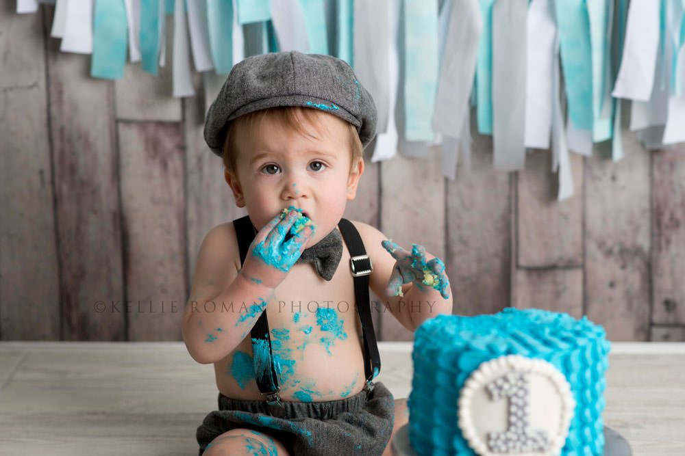 simple cake smash a little boy in front of a distressed wood backdrop digging into a teal colored cake with the number 1 on it he has teal frosting all over his mouth and chest