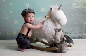 simple cake smash a little boy wearing a vintage outfit with suspenders holding onto a white fuzzy unicorn rocker a grey stuffed dragon is also in the shot he's in front of a teal backdrop with stars