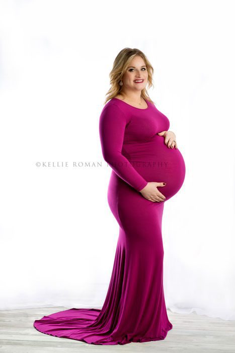 maternity photos a women wearing a pink gown who is pregnant looking at camera while holding her belly she's in front of a bright white backdrop