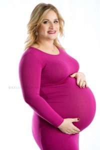 maternity pics a pregnant women standing in a bright pink gown in front of a white backdrop holding onto belly she's smiling at the camera