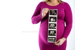 maternity photos a pregnant women wearing a tight pink gown holding strip of ultrasound photos only her middle section is in the photo and she's infant of a bright white backdrop