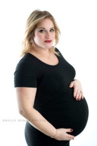 maternity photos women with pregnant belly in a black dress holding onto belly she's in a studio with a white backdrop