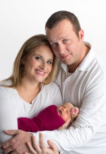 newborn girl in studio parents in milwaukee with their baby girl wrapped in a pink fabric swaddle mom is holding baby newborn and both her and dad are looking at camera