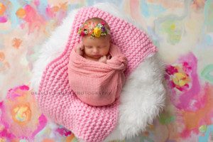 newborn girl in studio a newborn baby girl in milwaukee photo studio she's wrapped in a pink swaddle laying in a bucket on top of pink and white layering fabric there is a pink and colorful backdrop underneath her she has a colorful headband on