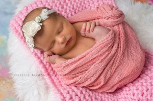 newborn girl in studio a newborn baby in milwaukee photo studio she's wrapped in a bright pink fabric laying on top of other layers of fabric she's holding onto one of her feet wearing a white headband