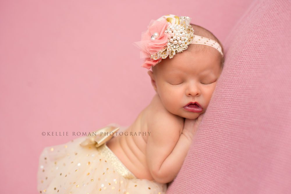 newborn girl in studio an infant girl laying asleep on a pink blanket she's wearing a gold tutu with a bow and a pink and gold headband she's in a milwaukee photographers studio
