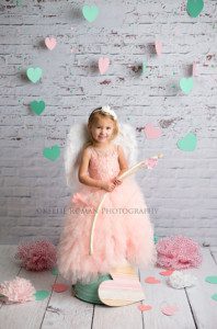 valentine mini session a toddler girl wearing a big puffy pink dress standing on a teal bucket she's holding a bow and arrow and is surrounded by hearts and pom poms
