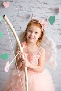 valentine mini session photos in milwaukee Wisconsin studio a girl wearing pink fluffy dress is holding a bow and arrow she's smiling at the camera and there are teal hearts behind her