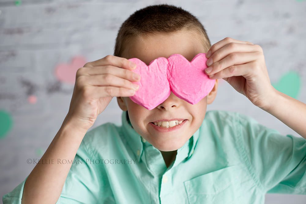 valentine mini session a boy with a teal button up shirt in milwaukee studio is holding two pink heart shaped candies in front of his eyes he's smiling with his mouth