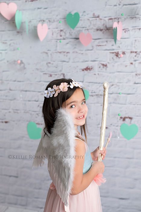 valentine mini session a girl wearing angel wings and holding a bow and arrow is looking at the camera teal hears are hanging in the backdrop and she has a floral crown on her head