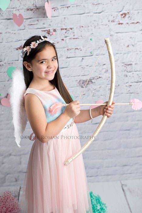 valentine mini a girl with a pink and blue dress on is wearing angel wings and holding a cupid bow and arrow she's in front of a white brick backdrop and has long brown hair she's smiling at the camera