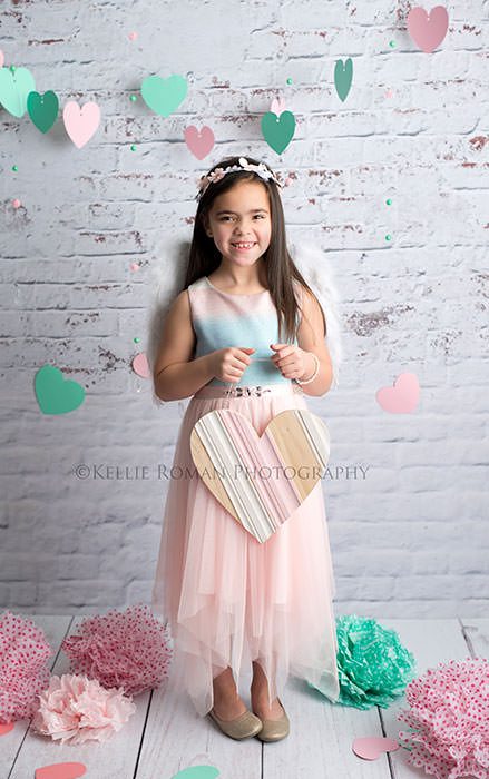 valentine mini session in milwaukee photographers studio a girl wearing a pink and blue dress is holding a pink wood heart she is looking at the camera and there are tissue paper pom poms on the floor around her