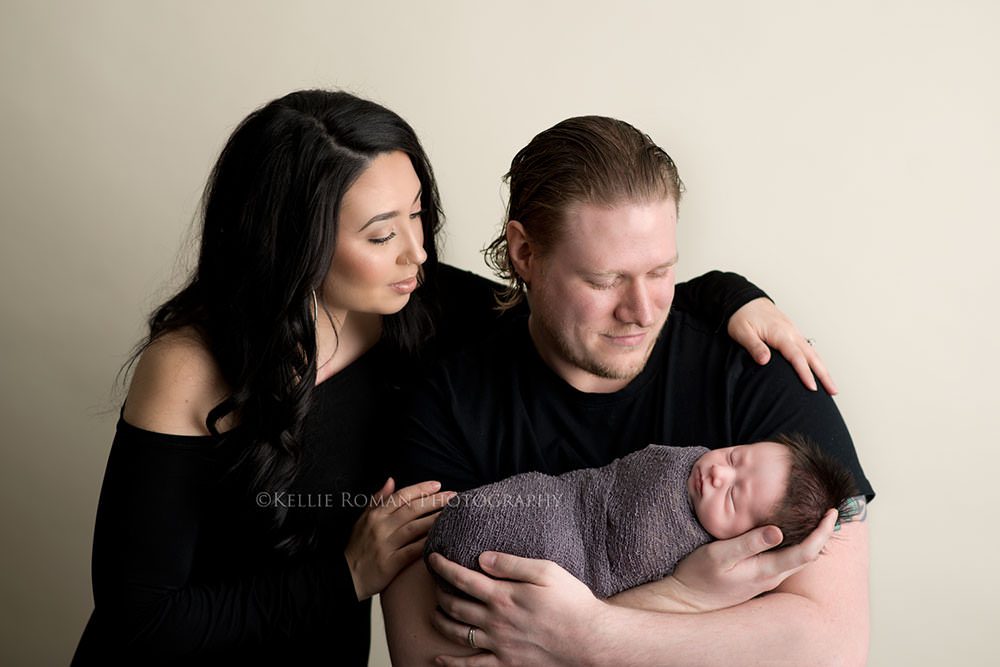 milwaukee newborn pics a husband and wife who are standing in milwaukee Wisconsin photographers studio wife has her arms around husbands shoulders and husband is holding newborn son who is sleeping the parents are both looking down at their new son