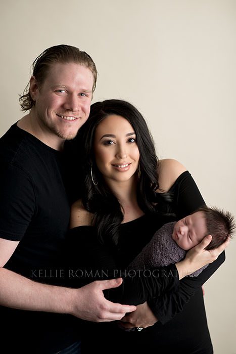 milwaukee newborn pics a family who is from kenosha having newborn pics taken in milwaukee photographers studio a mother and father are standing together while looking at the camera the mother is holding their new infant son who has lots of dark hair and is in a grey swaddle wrapped up sleeping
