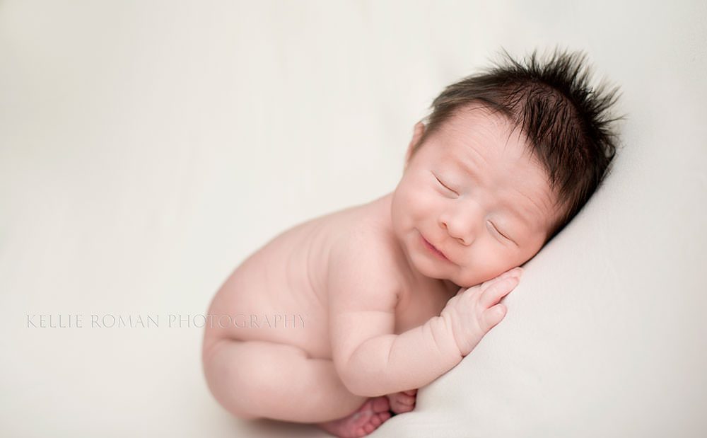 milwaukee newborn pics newborn boy who's is naked is posed on his stomach with his hands under head and his toes sticking out he has a lot of dark brown hair and is smiling
