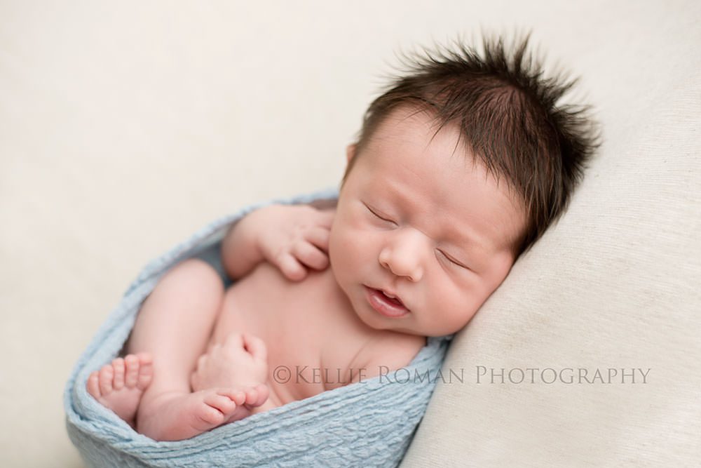 milwaukee newborn pics a newborn boy in milwaukee photo studio laying wrapped up on his back in a blue wrap he's on top of an ivory blanket and has his hands and feet curled up