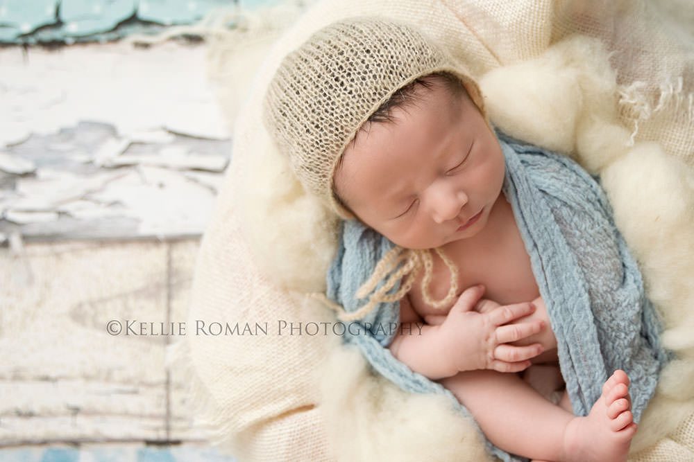 milwaukee newborn pics a newborn baby boy from kenosha Wisconsin in Milwaukee photo studio he's on top of a blue and ivory wood backdrop he's in a bucket wrapped in a blue wrap with an ivory bonnet on sleeping