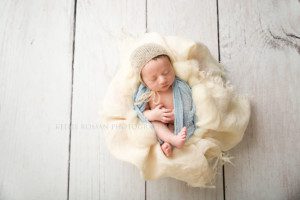 Milwaukee newborn pics a newborn from kenosha in Milwaukee Wisconsin photographers studio he's wrapped in a baby blue swaddle fabric with a tan bonnet on and is in a bucket with ivory fabric he's sleeping with hands folded