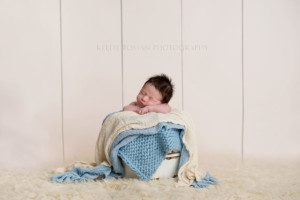 milwaukee newborn pics a newborn boy posed upright in a white bucket in front of an ivory wood backdrop the bucket has shades of blue and ivory fabric layering on top the newborn has lots of black hair he's in a Milwaukee photographers studio