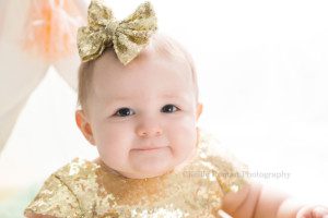 pastel milestone session a one year old toddler girl wearing a gold dress looking right into the lens of the camera she has a big gold sequin bow on her head