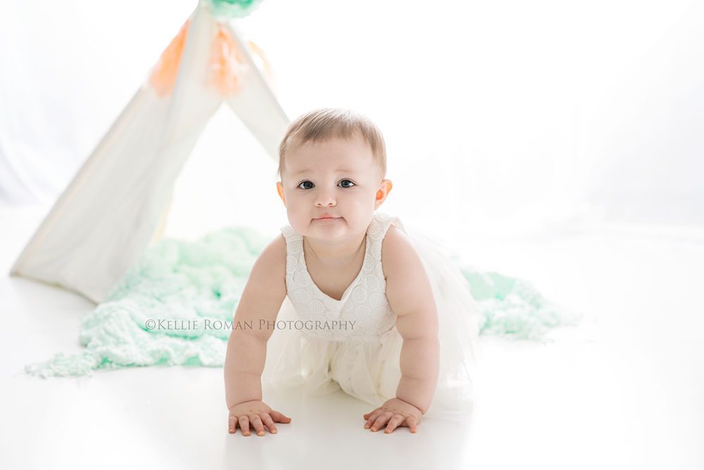 pastel milestone session one year old girl from Illinois who's getting her pics done by Milwaukee Wisconsin photographer she's on her hands and knees looking into the lens and there is a white teepee with flowers and teal blanket behind her it's very bright white because the pics is backlit