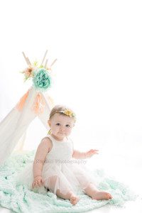 pastel milestone a one year old baby girl sitting on a teal blanket in front of a white teepee with flowers on it she's wearing a white dress and is looking at the camera the image is very bright white and backlit