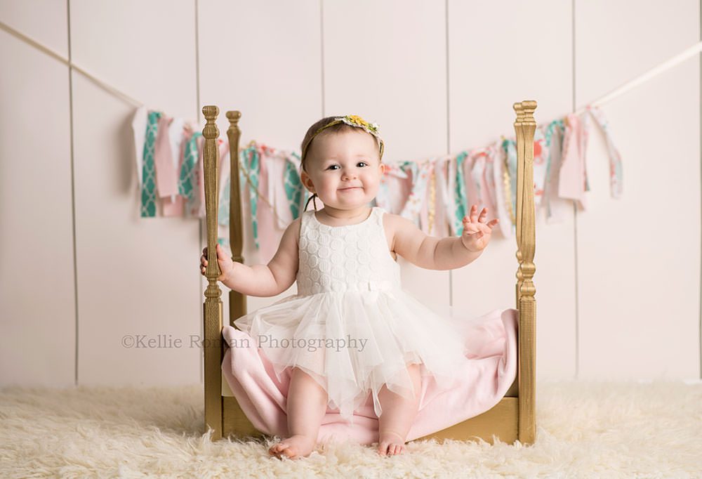 pastel milestone session one year old girl sitting on a gold bed in Milwaukee Wisconsin photography studio she's wearing a white dress and the bed is on a fuzzy ivory rug the backdrop is ivory wood with a pastel colored banner