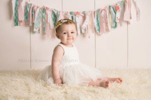 pastel milestone session one year old girl having pics taken in Milwaukee Wisconsin studio she's from Illinois she's sitting on an ivory fuzzy rug with an ivory wood backdrop she has a white dress on with a headband and there is a pastel colored banner behind her