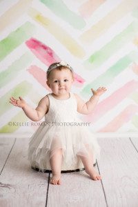 pastel milestone session one year old girl sitting on a white tub in a Milwaukee photography studio she has a white tutu dress on and has her hands out to the sides she's sitting in front of a pastel chevon backdrop