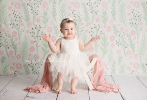 pastel milestone session one year old girl from Illinois in Milwaukee Wisconsin photographers studio she's wearing a white dress and is sitting on a white bench with a pink scarf over it the backdrop is pink and green floral and she has her hands sticking out to the side