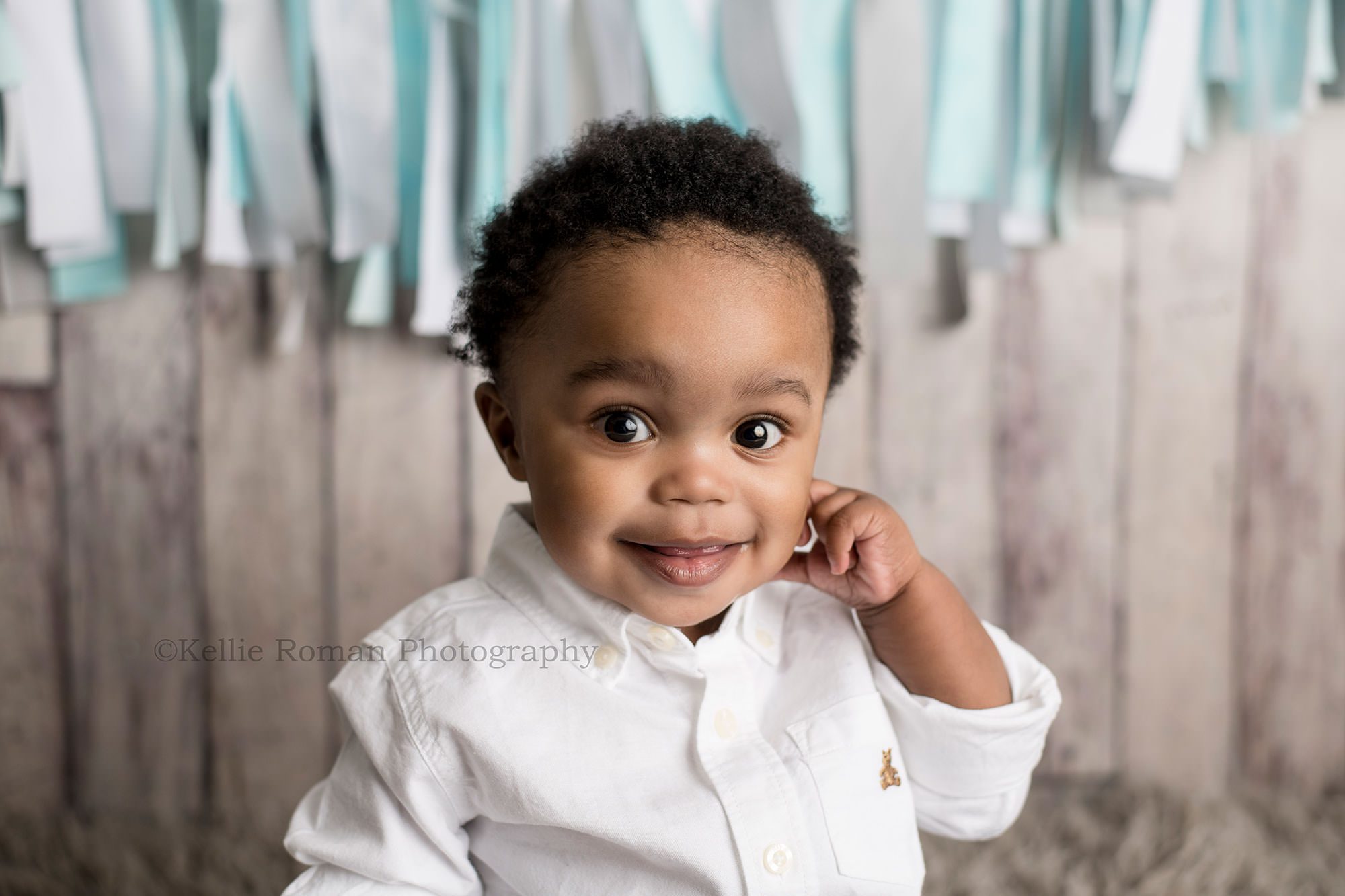 shades of blue cake smash a one year old boy having pics taken for his milestone session in Milwaukee Wisconsin photography studio he's wearing a white button up shirt with a wood backdrop and a fabric banner