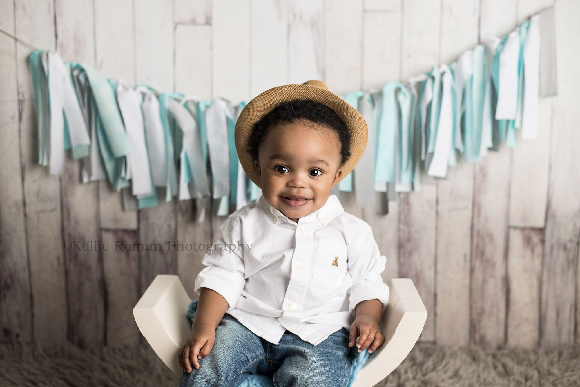 shades of blue cake smash one year old boy in a photography studio in Milwaukee sitting on a white curved bench he's wearing jeans and a white button up shirt with a hat smiling at the camera the backdrop is fake wood with a blue banner