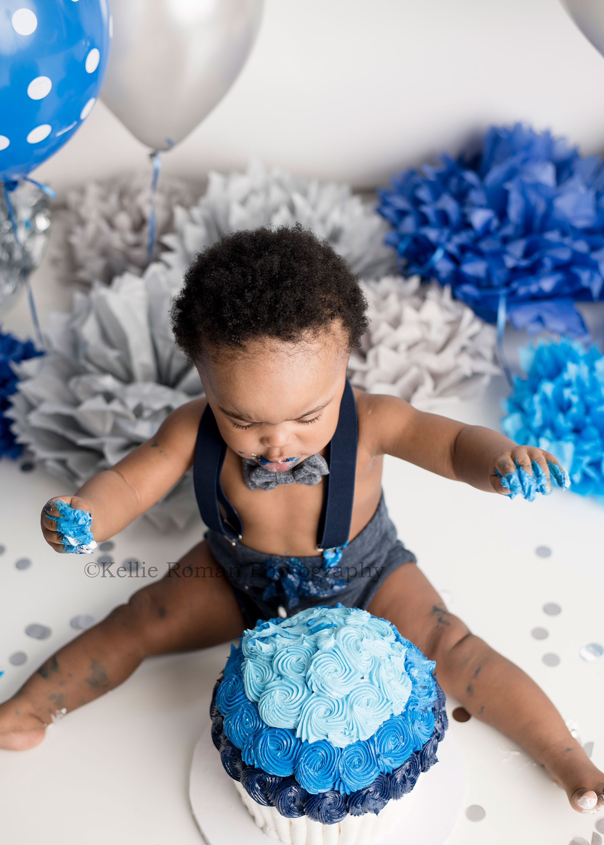 shades of blue cake smash one year old boy in a Milwaukee Wisconsins photographers studio he's sitting behind a blue and white back and has suspenders and a bow tie on he's surrounded by blue balloons