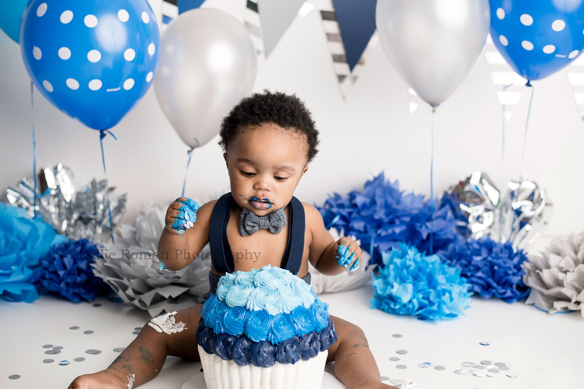 shades of blue cake smash one year old boy in a Milwaukee photography studio with a white and blue cake in front of him he's looking down with blue frosting on his legs and hands and has a white backdrop with blue balloons he's wearing a bow tie and suspenders