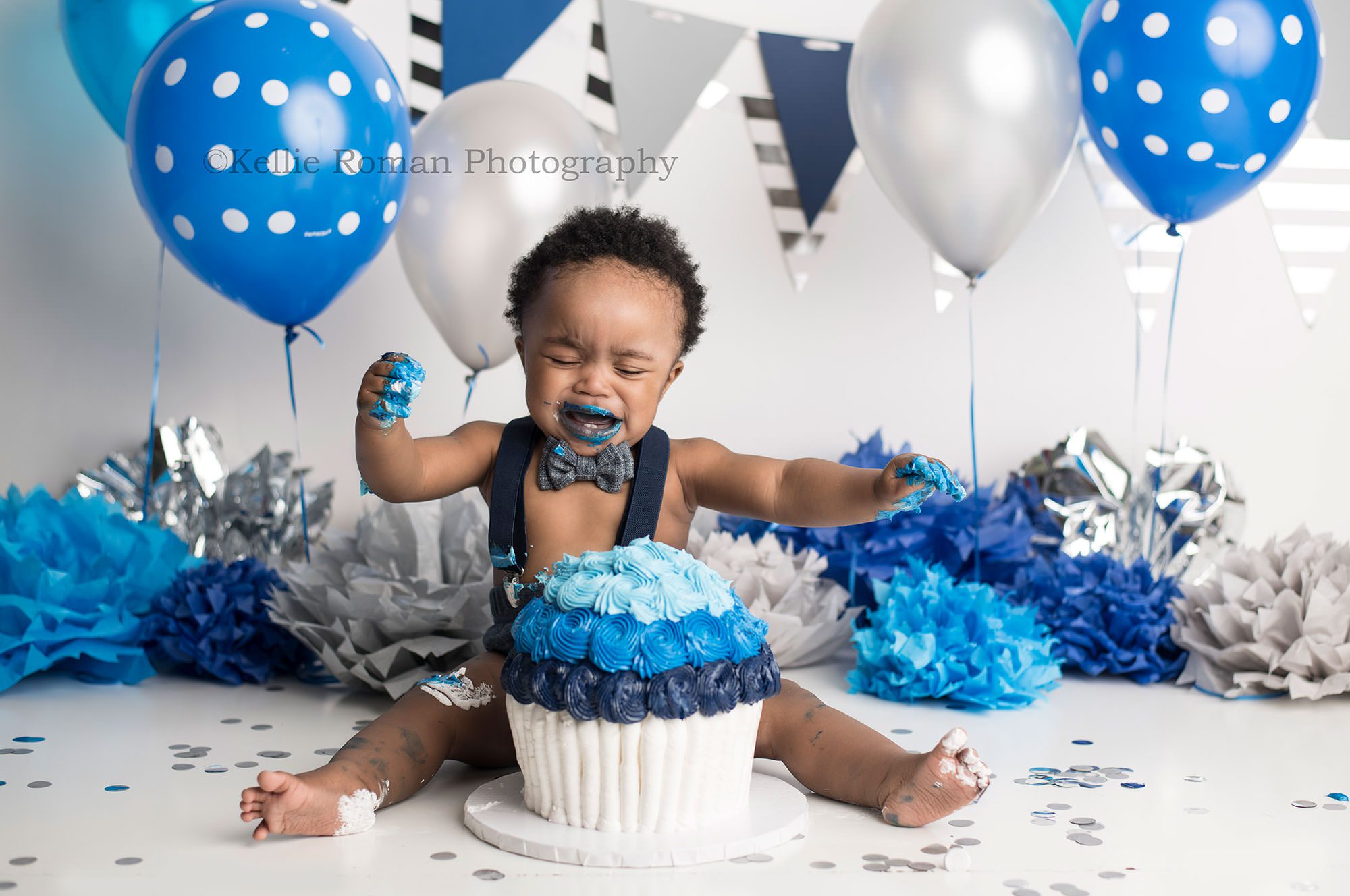 shades of blue cake smash one year old boy in Milwaukee Wisconsin photographers studio having pics taken wit ha blue and white large cake the backdrop is filled with shades of blue balloons and tissue paper pom poms. the boy is wearing blue suspenders and a bow tie and is crying with frosting on his feet and hands