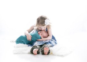 texas roots a newborn session with a kenosha family in Milwaukee Wisconsin photography studio a three year old girl is sitting with her legs out and is holding her swaddled newborn brother on top of a white rug they are in shades of blue